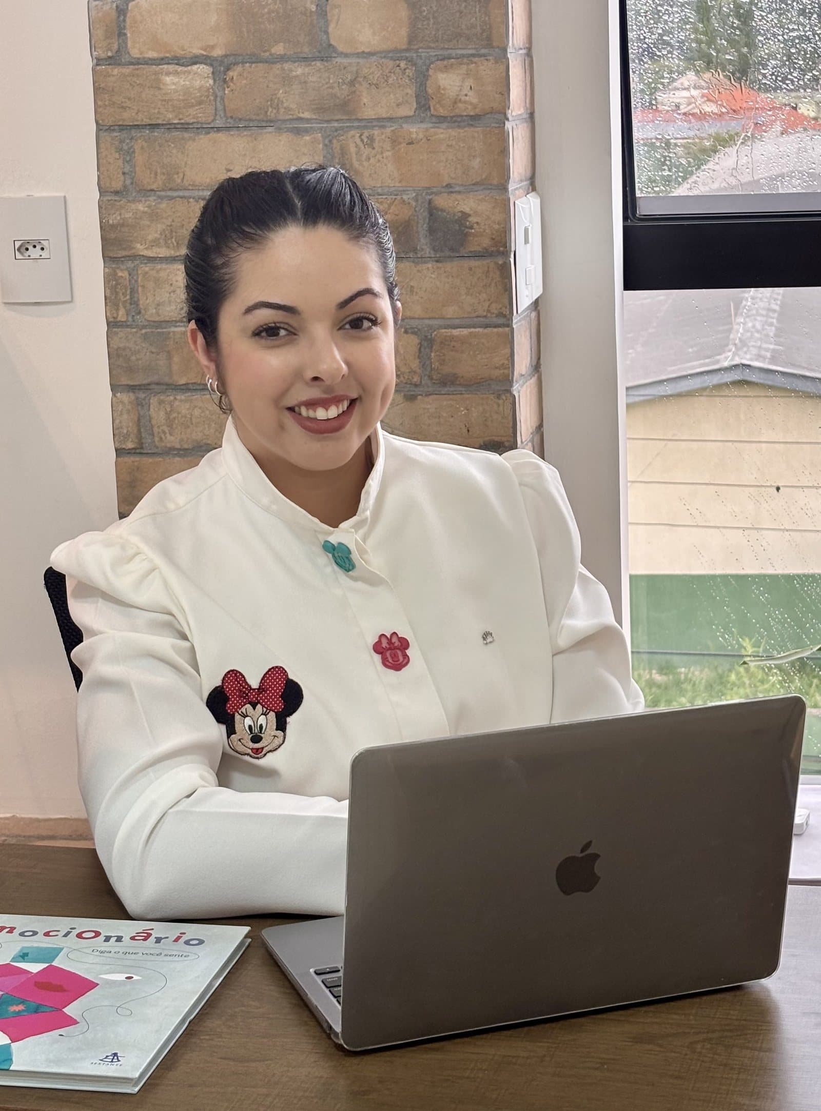 Maria Izabel Rubens sentada à mesa do consultório, de blusa branca, com um notebook à frente.
